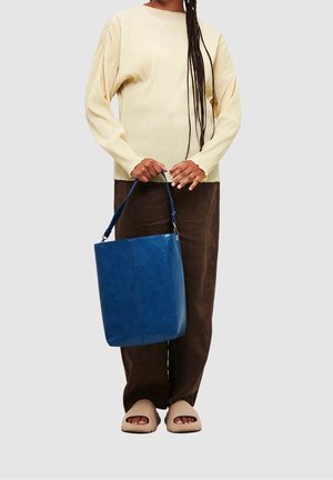 Personne portant un pull beige et un pantalon marron, tenant un grand sac à main en cuir bleu, et portant des chaussures ouvertes beiges.