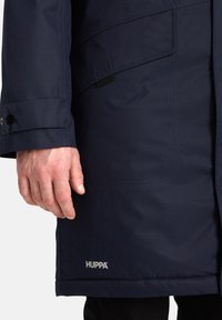 Cappotto impermeabile blu navy con finitura testurizzata, dotato di tasche frontali, polsini con bottoni e un'etichetta del logo all'orlo.