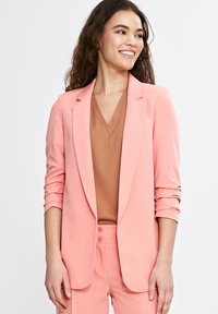 Bladoróżowa marynarka o dopasowanym kroju, z klapami i podwiniętymi rękawami. Brązowy top w serek pod spodem. Gładki materiał z minimalnymi detalami.
