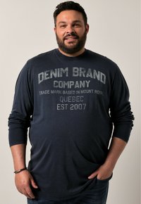 Námořnicky modrá dlouhá rukávová košile s kulatým výstřihem, s lehce šedým textem: "DENIM BRAND COMPANY, QUEBEC EST 2007." Hladká látka, volný střih.