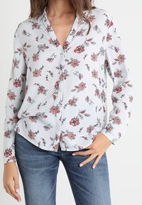 Chemise à boutons en tissu léger, avec un imprimé floral rouge et vert sur un fond rayé clair. Manches longues et coupe décontractée.