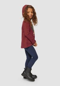 Bordeaux hoodie met een grafische print, gecombineerd met marineblauwe leggings en zwarte veterschoenen. Krullend haar, staande pose, lichtgrijze achtergrond.