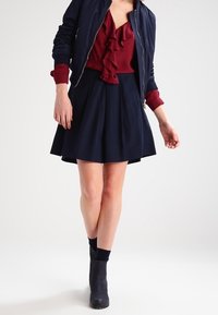 Donna che indossa un giubbotto blu navy, una blusa bordeaux con volants, una gonna plissettata blu navy e stivaletti neri con tacco, in piedi su uno sfondo bianco.