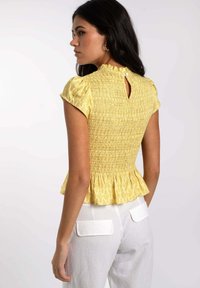 Top amarillo con smock, mangas cortas abullonadas y un escote con volantes. Presenta un detalle de abertura en la parte posterior y un dobladillo de peplum. Combinado con pantalones blancos.