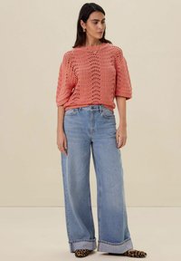 Koraalroze gebreide trui met open structuur, brede korte mouwen. Hoge taille lichtblauwe wijde jeans en zwarte laarsjes met luipaardprint.