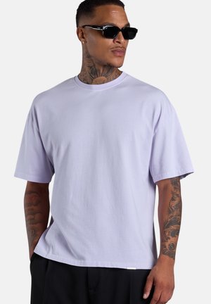 Mann mit Hals- und Armtattoos, der schwarze Sonnenbrillen, ein hellviolettes übergroßes T-Shirt und schwarze Hosen trägt und nach rechts schaut.