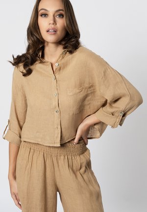 Donna che indossa una camicia corta in lino beige con bottoni e maniche arrotolate e pantaloni raccolti a vita alta abbinati, mano sul fianco contro uno sfondo semplice.