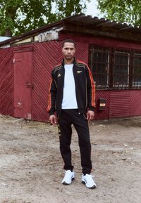 Ensemble de survêtement noir avec des rayures orange, comprenant une veste zippée et un pantalon ajusté. T-shirt blanc en dessous, complété par des chaussures de sport blanches.