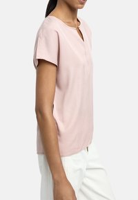 Jasnoróżowa bluzka z krótkim rękawem o okrągłym dekolcie i kluczykowym wycięciem z przodu, wykonana z gładkiego, lekkiego materiału.