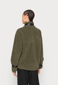 Pullover in fleece verde oliva con colletto alto e dettagli neri sui polsini. Tessuto morbido con vestibilità rilassata e cuciture dettagliate sul retro.