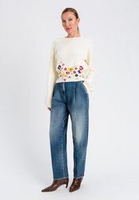 Maglione a costine crema con ricami floreali in vita, abbinato a pantaloni in denim blu a vita alta e tacchi a punta marroni.