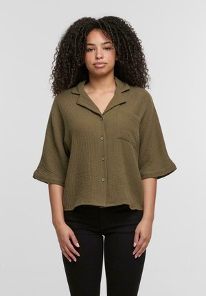 Camicia verde oliva con bottoni e tasca, realizzata in tessuto testurizzato e caratterizzata da un colletto e maniche ampie, abbinata a pantaloni neri.