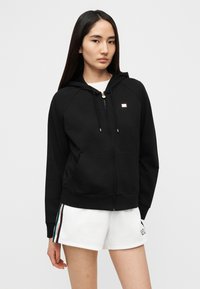 Μαύρη ζακέτα με φερμουάρ, με μπροστινή τσέπη, ραγκλάν μανίκια, κουκούλα με κορδόνι και μικρό λογότυπο στο στήθος, συνδυασμένη με λευκά σορτς.