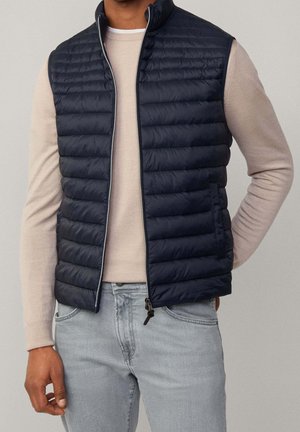 Homme portant une veste matelassée noire sur une chemise beige à manches longues et un jean gris clair, debout avec une main dans la poche.