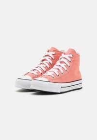 Zapatillas altas de lona de color coral. Presentan cordones blancos, ojetes plateados y una puntera de goma blanca con suela a rayas negras.