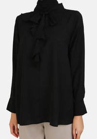 Black blouse with a ruffled front, bow tie detail at the collar, long sleeves, and a smooth, flowing fabric. Paired with light-colored trousers.