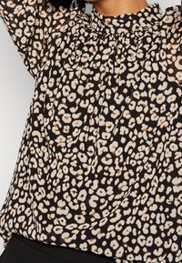 Blouse noire avec un motif léopard en crème et marron, comportant des détails plissés et un col haut. Tissu doux et léger.