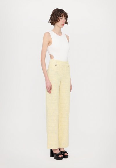 Valkoinen hihaton bodysuit, jossa on leikkuuyksityiskohtia, yhdistettynä korkeavyötäröisiin keltaisiin tweed-legginsseihin, joissa on korostusnapit ja teksturoitu kangas.