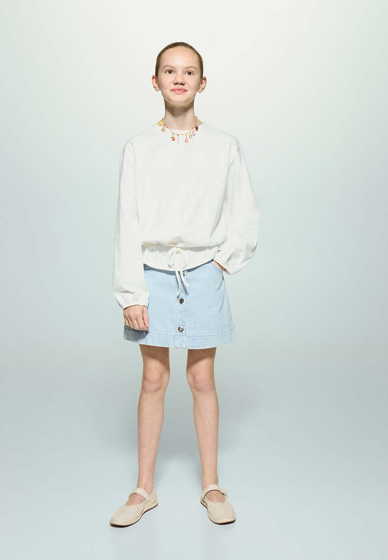 Jeune fille debout, une main dans la poche, portant un haut à manches longues blanc, une jupe bleu clair boutonnée, des chaussures beiges et un collier coloré.