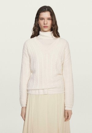 Maglione a trecce di colore crema chiaro con scollo a V e maniche lunghe, indossato su un dolcevita bianco. Texture morbida con polsini e orlo a coste.
