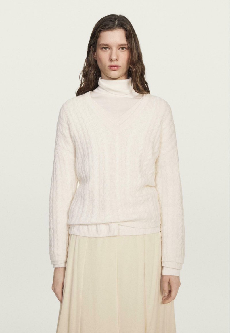 Maglione a trecce di colore crema chiaro con scollo a V e maniche lunghe, indossato su un dolcevita bianco. Texture morbida con polsini e orlo a coste.