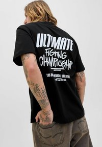 Persona con cabello largo vistiendo una camiseta negra de "Ultimate Fighting Championship" y pantalones marrones con múltiples tatuajes en los brazos, girado ligeramente hacia un lado.