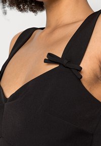 Robe noire à larges bretelles, dotée d'un décolleté en V et d'un nœud décoratif sur la bretelle gauche. Texture lisse et design simple avec peu de détails.