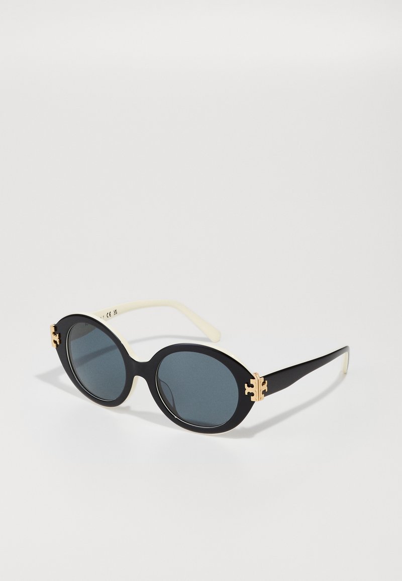 Ochelari de soare ovali negri cu lentile întunecate, brate de culoare crem și accente din metal aurit pe față. Suprafață netedă, design modern.