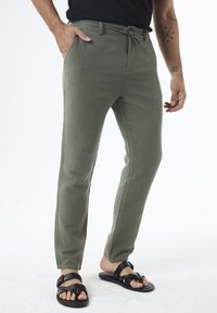 Lightweight, olive-green pants with a drawstring waist, side pockets, and a tapered fit, paired with black sandals. Textured fabric.