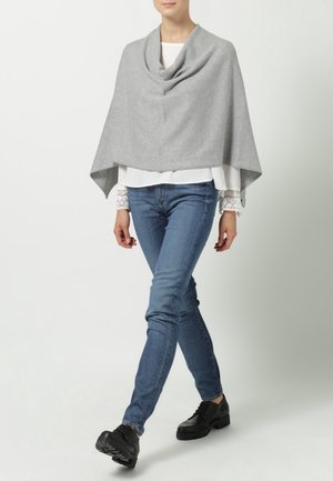 Femme portant un poncho gris sur un chemisier blanc avec des poignets en dentelle, un jean bleu et des chaussures noires à lacets, marchant devant un fond uni.