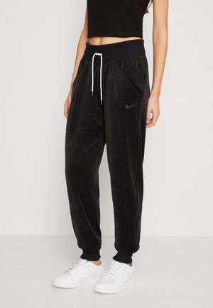 Kobieta w czarnych spodniach dresowych Nike ze sznurkiem, o fakturowanym materiale i białych trampkach, stoi na jednolitym jasnym tle.