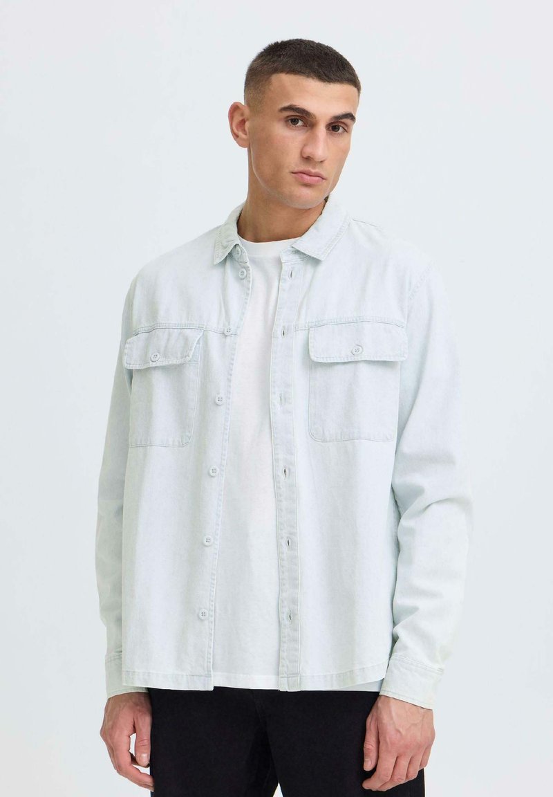 Jeune homme aux cheveux courts et foncés portant une chemise en jean claire boutonnée par-dessus un t-shirt blanc, sur un fond uni.