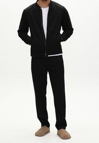 Mann trägt eine schwarze Zip-up-Jacke, schwarze Hose, weißes T-Shirt und braune Slipper, steht mit den Händen in den Taschen vor weißem Hintergrund.