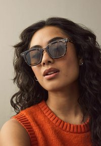 Ochelari de soare maro cu model țestoasă, cu rame pătrate, lentile gri și accente argintii. Purtați cu un crop top portocaliu fără mâneci.