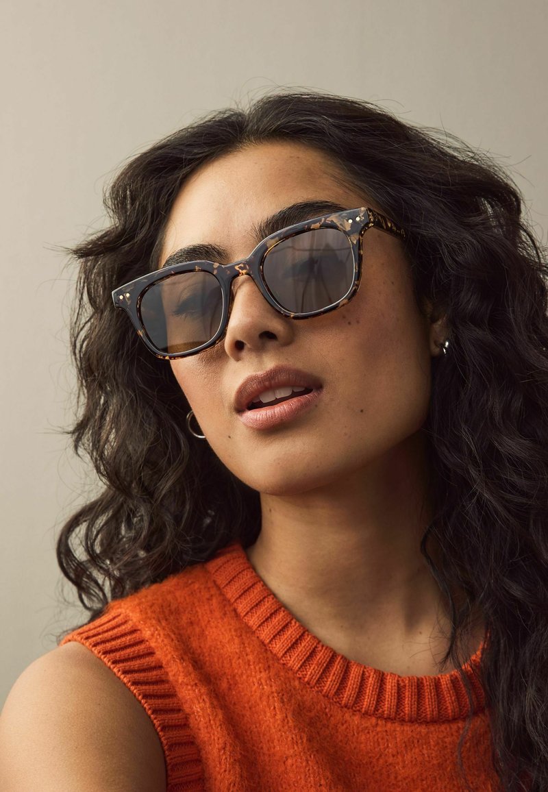 Lunettes de soleil en écaille brune avec des montures carrées, des verres gris et des accents argentés. Portées avec un pull sans manches orange.