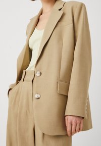 Béžový oblekový blazer se dvěma knoflíky, klopovými límci a přední kapsou. Texturovaná tkanina; spárováno s lehkým žebrovaným topem.
