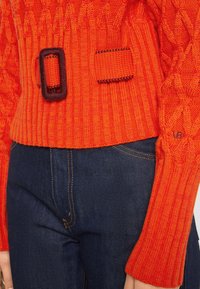 Pull en laine orange avec texture côtelée, agrémenté d'une ceinture marron. AssorTt avec un denim bleu foncé. Vue en gros plan de la zone du torse.