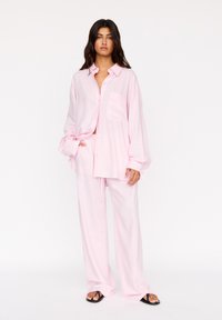 Lichtroze linnen overhemd en bijpassende wijde broek. Het overhemd heeft een zak en knopen; de losse pasvorm biedt comfort. Zwarte slippers maken de look compleet.