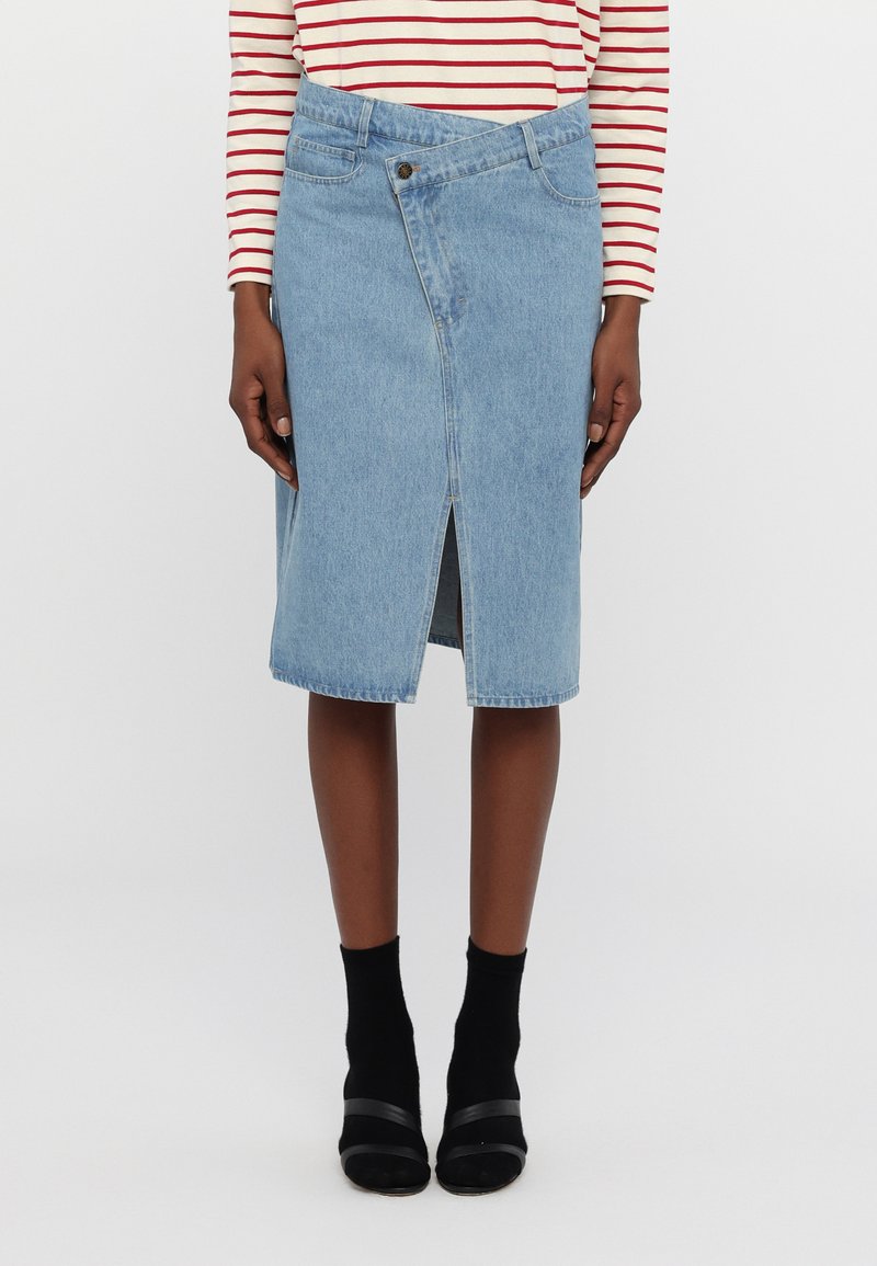 Denim krilo z visokim razporkom, svetlo modre barve in klasičnega kroja s sprednjim gumbom. Obuto z črnimi gležnjarji in petami.