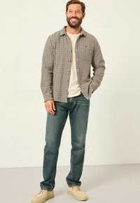 Braun- und beigetarnierte Baumwollhemd mit Knöpfen, kombiniert mit hellblauen Jeans und beigen Schuhen. Der Hintergrund ist in einer einfarbigen, hellen Farbe.