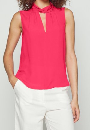 Femme portant un chemisier rose vif sans manches avec un col en trou de serrure et un pantalon blanc, debout avec une main dans la poche.