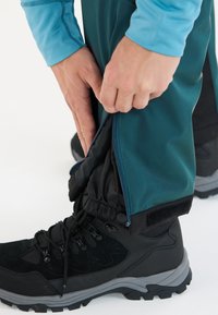 Hände stellen den Reißverschluss und den Klettverschluss an türkisfarbenen Outdoor-Hosen über schwarzen Wanderschuhen mit grauen Sohlen auf einem weißen Hintergrund ein.