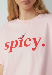 Helles rosa Baumwoll-T-Shirt mit rotem Text, der "spicy" sagt. Eine kleine rote Chili-Pfeffer-Akzent befindet sich über dem Buchstaben 's.'