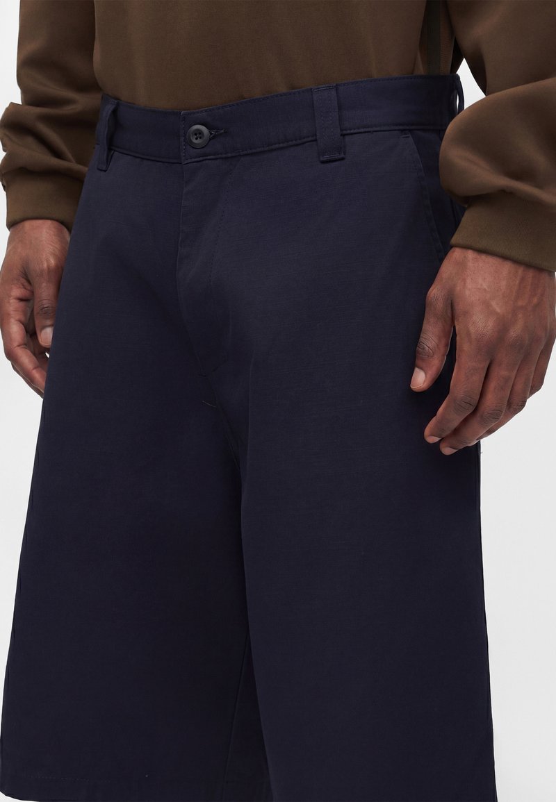 Mand iført mørkeblå shorts og en brun langærmet skjorte, med afslappede hænder ved siderne, beskåret ved talje og lår.
