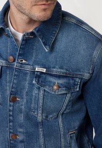 Donkerblauwe spijkerjas met een klassieke snit, voorzien van een kraag, borstzakken en bruin metalen knopen. Merklabel zichtbaar.