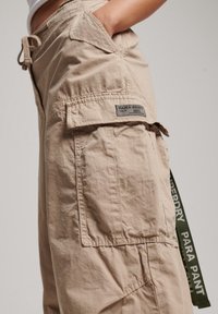 Beige cargobyxor i lätt tyg, med dragsko i midjan, sidofickor och en etikettpatch på låret. Accentueras med olivgrön logotyp.