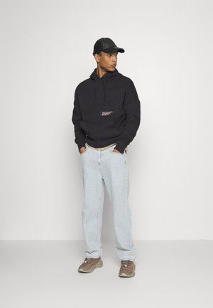 Felpa con cappuccio nera con logo frontale, jeans azzurri chiari e sneaker marroni. Il modello indossa un berretto nero. Outfit semplice e casual.