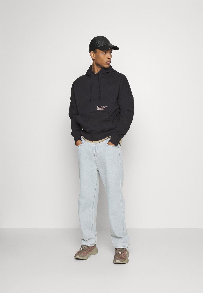 Felpa con cappuccio nera con logo frontale, jeans azzurri chiari e sneaker marroni. Il modello indossa un berretto nero. Outfit semplice e casual.