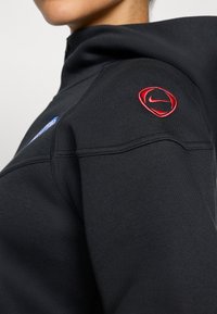 Svart hettegenser med høyt krage, med en rød Nike-logo på skulderen og et blått grafisk design på framsiden. Glatt tekstur.