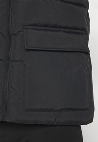 Black puffer jacket featuring a zip closure, textured fabric, and a large patch pocket on the front. The material appears lightweight and insulated.